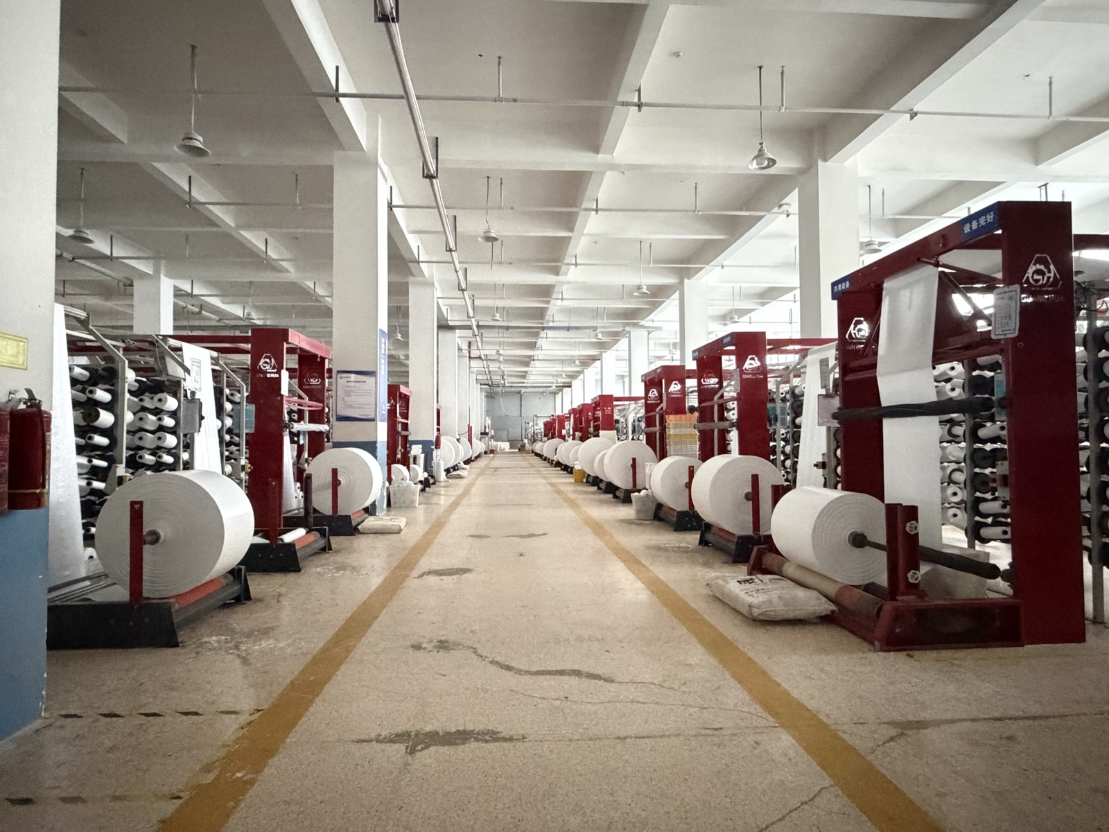 Two rows of circular looms with bobbin creels and woven fabric rolls