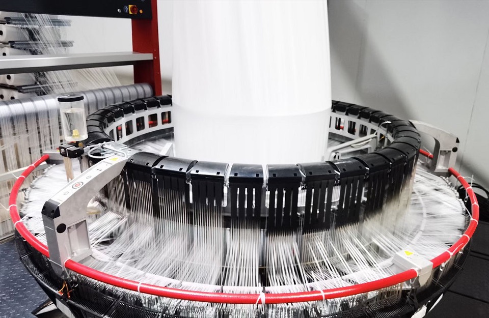 Circular loom in operation, tape yarns feeding from the creel into a woven fabric tube rising through the centre