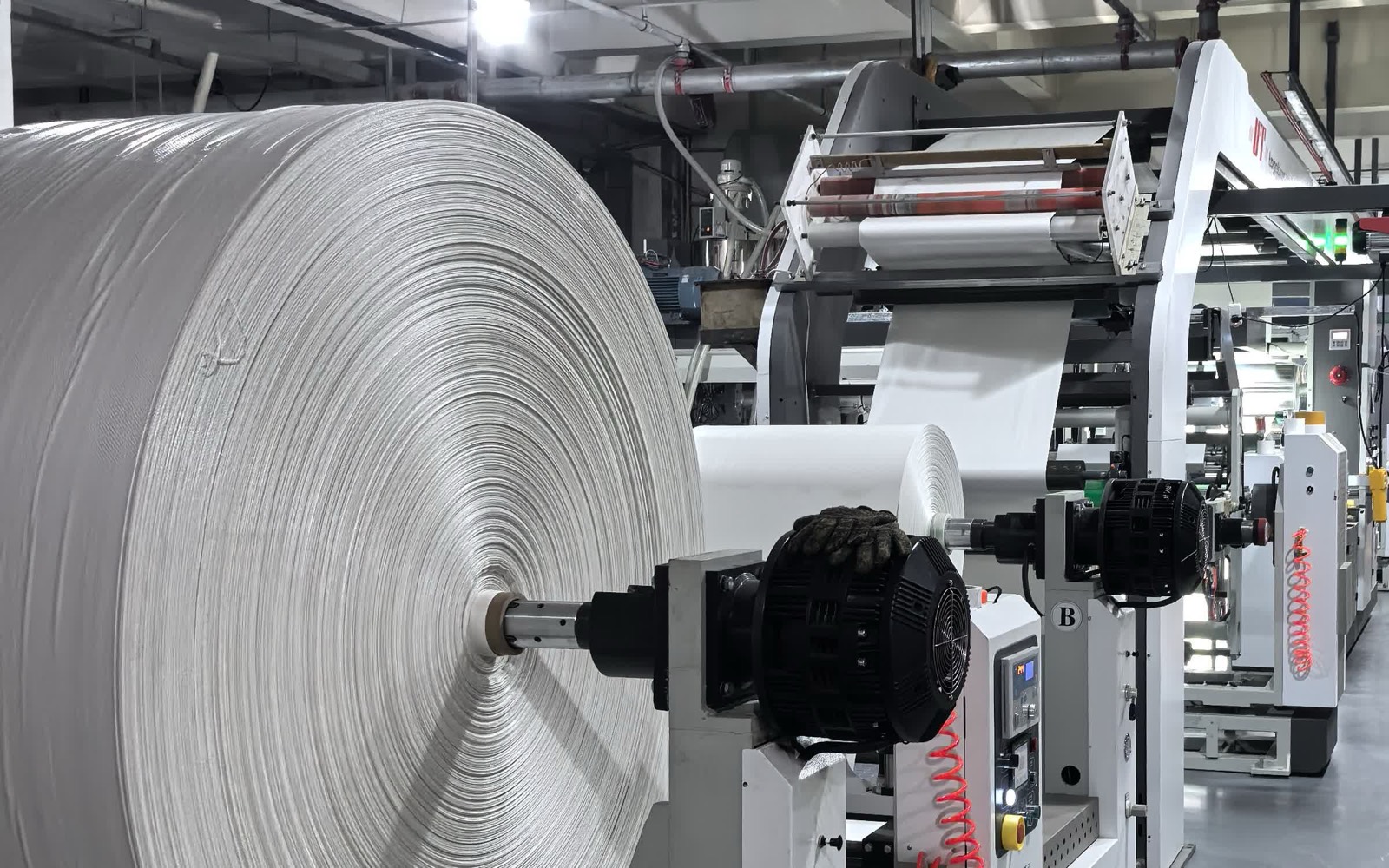 Large white woven fabric roll on a laminating line unwind stand, the web running up over the rollers into the line